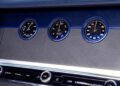 Close-up of three circular dials on a car dashboard, displaying time, temperature, and compass direction, set against a sleek blue and silver panel in true Bentley style.