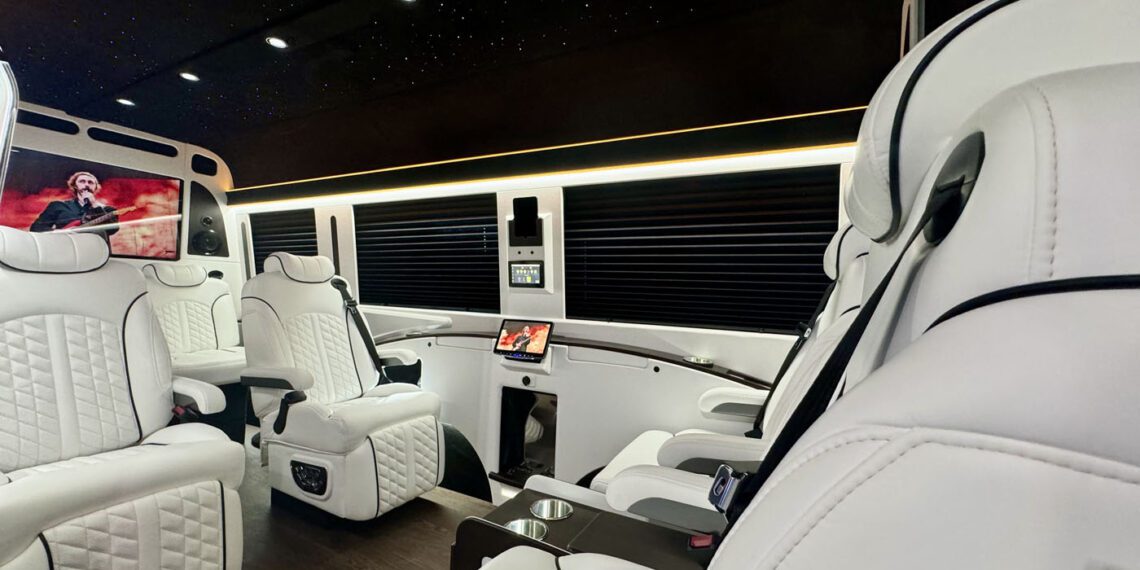 The 2024 Jet Sprinter by First Class Customs features a lavish interior with white leather seats, wood flooring, ambient lighting, and a ceiling that mimics a starry night. A sleek TV screen graces the background, creating an atmosphere of pure luxury.