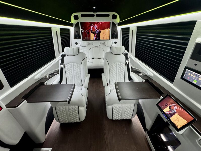 The 2024 Jet Sprinter exudes elegance with its luxury van interior, featuring reclining white leather seats, fold-out tray tables, and a television screen at the front. Designed by First Class Customs, it offers an entertainment control panel for an elite travel experience.