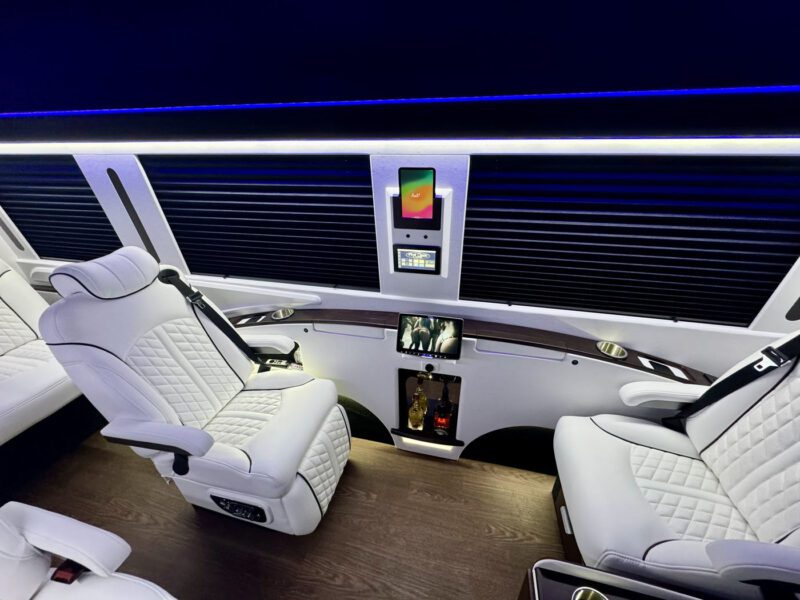 The Ultimate Class Jet Sprinter by First Class Customs offers a luxury van interior with plush white leather seats, elegant wood flooring, an overhead screen, and sophisticated bottle storage.