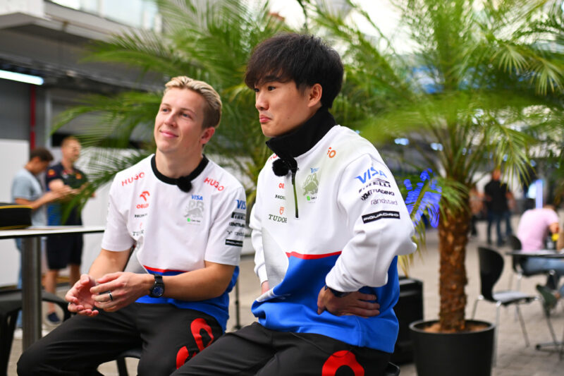 Two individuals sit together in branded team jackets, surrounded by potted plants in an outdoor setting. They're eagerly discussing the excitement of the upcoming 2025 F1 season and debating their favorite drivers' positions on the starting grid.