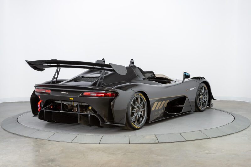 A sleek black sports car with the Flatrock Ultimate Package, combining speed and elegance, boasts a large rear spoiler and race-ready features. It's displayed on a circular platform in a showroom setting, captivating every onlooker.