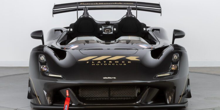 A black, sleek race car exuding speed and elegance with aerodynamic features and prominent logos on the hood and seats is showcased in a well-lit studio setting, offering the Flatrock Ultimate Package for an unforgettable $2 Million Experience.