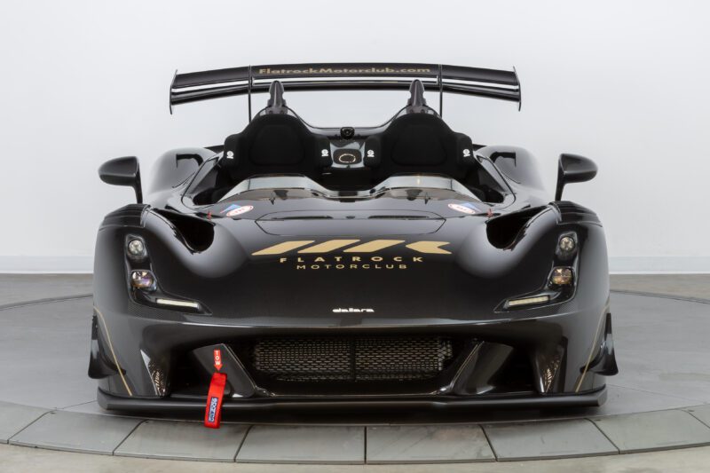 A black, sleek race car exuding speed and elegance with aerodynamic features and prominent logos on the hood and seats is showcased in a well-lit studio setting, offering the Flatrock Ultimate Package for an unforgettable $2 Million Experience.