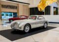 A vintage silver Aston Martin is displayed in a showroom at the Fontainebleau Las Vegas, alongside a red classic car, both on black platforms. This setting is part of an $18 million car collection that exudes elegance and modernity.