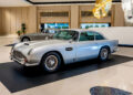 In a Las Vegas showroom, a silver vintage car is displayed among elegant décor, with an Aston Martin adding to the allure of this exquisite car collection in the background.