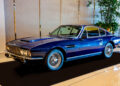 A classic blue Aston Martin sports car is displayed indoors, with lush plants in the background, evoking the allure of an exclusive Las Vegas car collection.