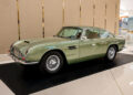 A classic green Aston Martin sits on display indoors in Las Vegas, elegantly parked on a black platform against a beige partition, adding timeless charm to the exquisite car collection.