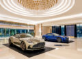 Two luxury sports cars, including a sleek Aston Martin, are showcased in an elegant Las Vegas showroom, illuminated by a large, ornate chandelier above and complemented by polished marble flooring.