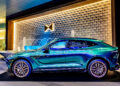 A teal luxury Aston Martin SUV is elegantly parked in front of a textured wall with a fountain, reminiscent of the opulence found at Fontainebleau Las Vegas.