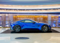 A sleek, blue Aston Martin sports car is displayed on a raised platform indoors, surrounded by modern architectural elements, as if it were the crown jewel of a Las Vegas car collection.