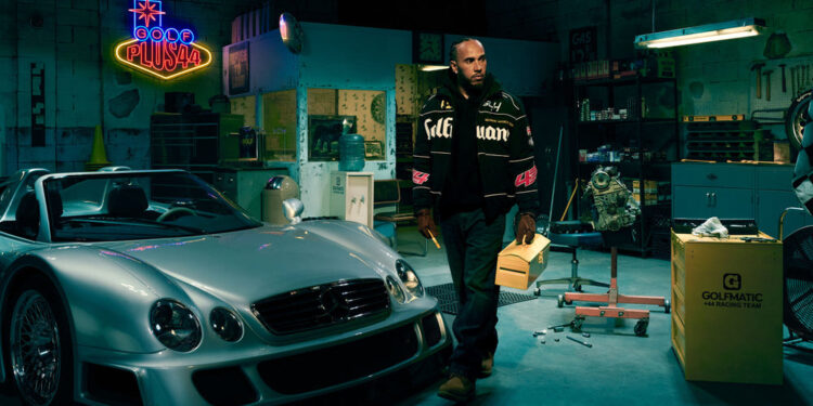 A man stands in a garage next to a silver sports car, holding a yellow folder. A neon sign reads "Gold Plush" in the background, reminiscent of the F1-Inspired Las Vegas Collection.