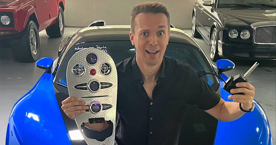 A person holds a car key and panel, sitting on a blue-and-black sports car in a garage filled with luxury cars, including a stunning 2008 Bugatti Veyron from Chicago Motor Cars.