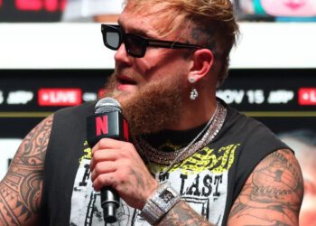 A man with sunglasses and tattoos holds a microphone, wearing a printed sleeveless shirt, multiple necklaces, and earrings. He sports a $7M watch while channeling Jake Paul's bold style.