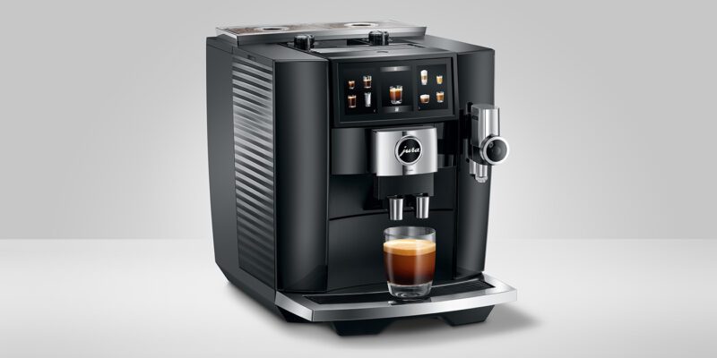 A black automatic espresso machine with a display panel elegantly brews a cup of espresso on a white surface, making it the perfect holiday gift for coffee aficionados who appreciate luxury machines.