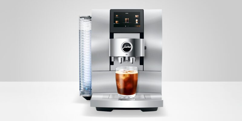 A silver coffee machine, perfect for coffee aficionados, brews iced coffee into a glass on a white surface—a luxury machine that's an ideal holiday gift.