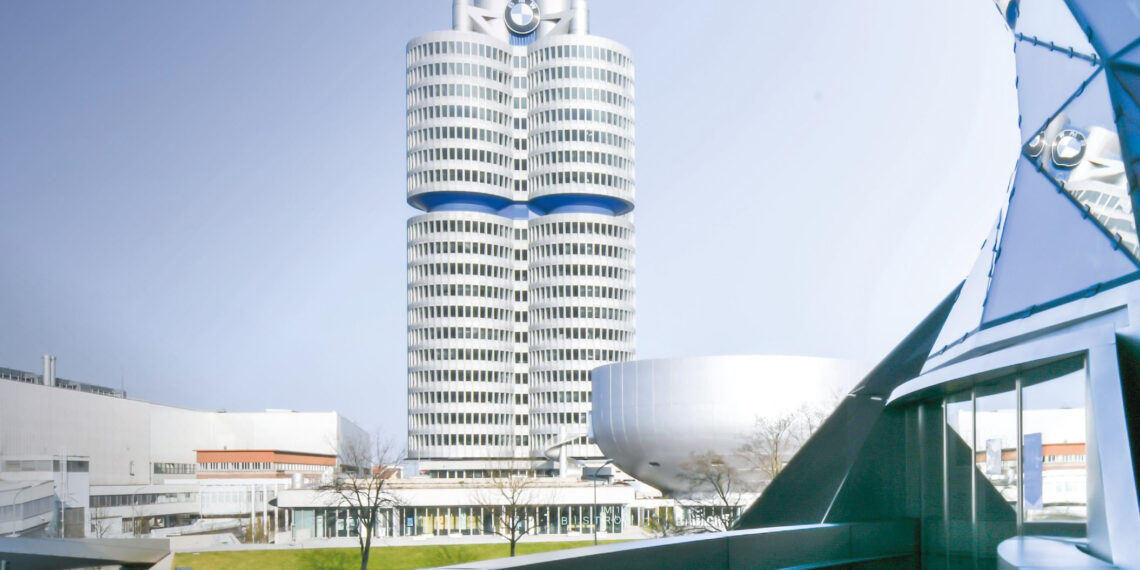 Set against a clear blue sky, the tall, cylindrical office building with a unique design and BMW Group logo on top stands proudly amidst a modern complex, symbolizing the impressive results anticipated in Q3 2024.