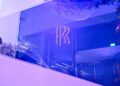 The elegant glass wall adorned with the Rolls-Royce logo and purple lighting creates a mesmerizing ambiance in the Abu Dhabi showroom, enhanced by lush foliage and sleek ceiling lights.