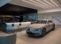 A luxury Rolls-Royce is elegantly displayed in a modern Abu Dhabi showroom, featuring a sleek design with a bar area adorned with teal chairs and decorative purple flowers.