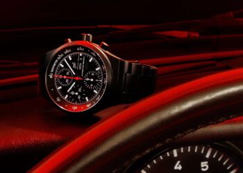A Porsche Design chronograph wristwatch with a red-accented bezel rests on a car dashboard, with the steering wheel and speedometer partially visible in the foreground.