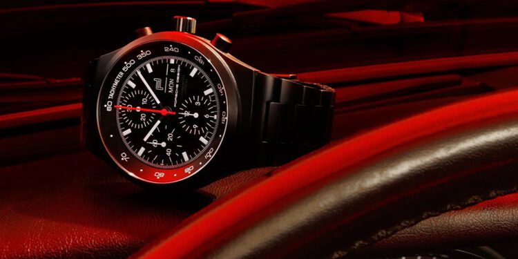 A Porsche Design chronograph wristwatch with a red-accented bezel rests on a car dashboard, with the steering wheel and speedometer partially visible in the foreground.