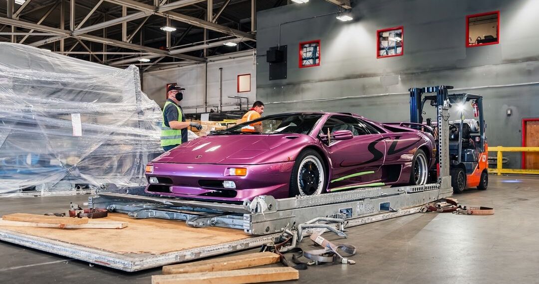 The Lamborghini Diablo SV in striking Viola 30th is positioned on a platform inside a CURATED warehouse. Nearby, two workers and a forklift stand ready to assist.