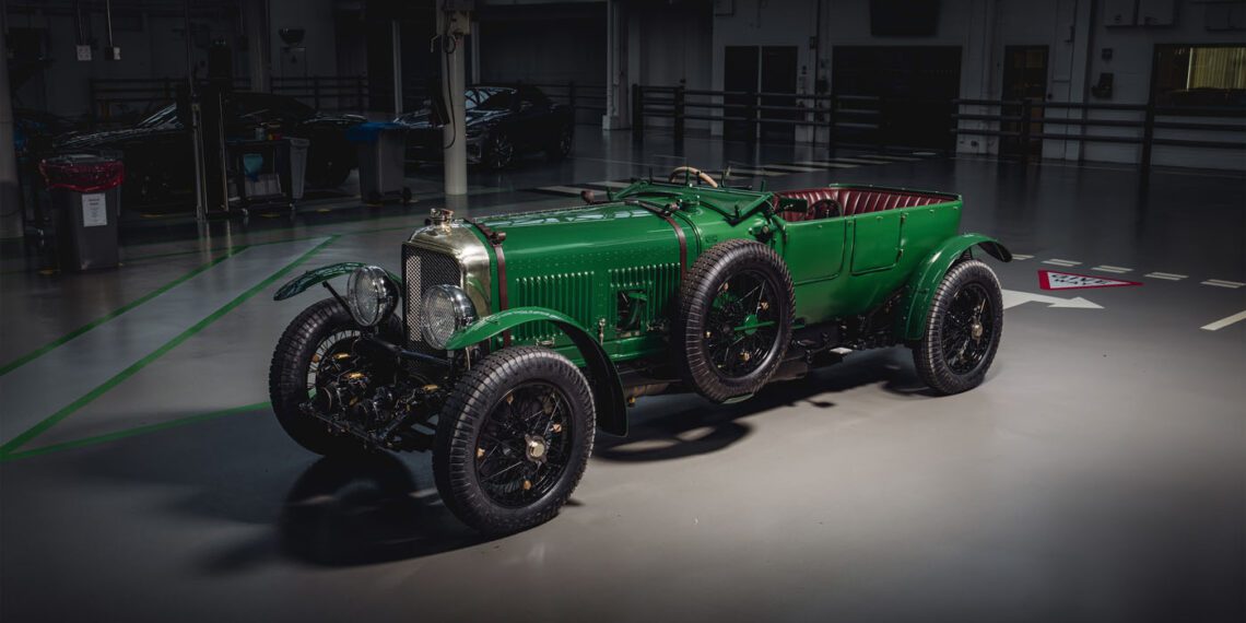 A vintage green Bentley, part of the Speed Six Continuation Series, stands parked indoors under dim lighting, showcasing classic design features and large spoked wheels with a luxurious red leather interior.