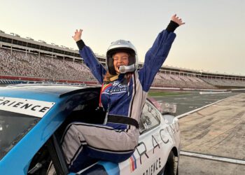 Person in a racing suit and helmet sits triumphantly on a NASCAR race car with arms raised in celebration at a racetrack, channeling the thrill of an unforgettable racing experience.