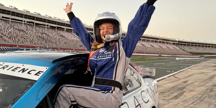 Person in a racing suit and helmet sits triumphantly on a NASCAR race car with arms raised in celebration at a racetrack, channeling the thrill of an unforgettable racing experience.