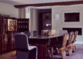 Nestled in The Mews, this modern dining room features a sleek wooden table with eight chairs, a display cabinet showcasing bottles, and a wall-mounted TV. An area rug graces the wooden floor beneath exposed beams, ensuring an exquisite client experience.