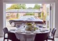 A round dining table set with white plates and glassware overlooks a parked Bentley outside, contrasting elegantly against the brick building of The Mews.