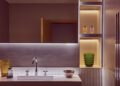 Modern bathroom with a sleek sink, chrome faucet, and decorative items including a green textured vase, colored glass, and metallic bowl on illuminated shelves—crafted to enhance the client experience reminiscent of The Mews elegance.