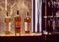 Three bottles of Macallan whiskey are displayed on a marble shelf at The Mews, flanked by shelves of glassware. A Bentley-emblazoned chair is partially visible in the foreground, enhancing the luxurious client experience.