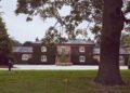 A large brick house with ivy-covered walls stands behind a tree in the foreground, exuding charm reminiscent of The Mews, and is surrounded by a manicured lawn.