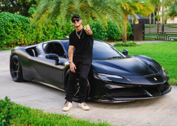 A person in black clothes and a cap sits confidently on a sleek Ferrari SF90 Stradale, giving a thumbs up. The powerful 986HP machine stands out against the lush greenery, capturing the vibe of WhistlinDiesel's adventurous spirit.