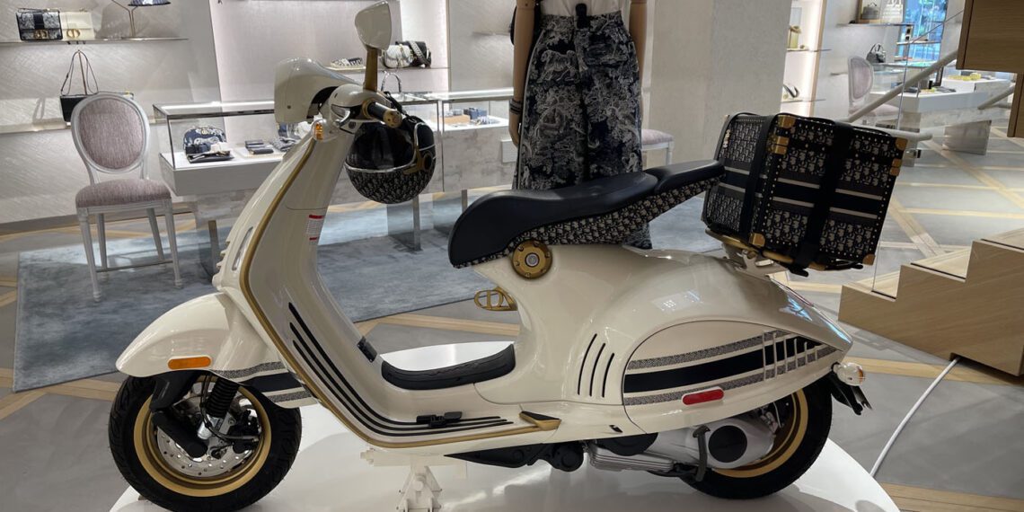 A white and black scooter with a patterned seat and storage box on display in a store. Shopping bags and other items are visible in the background.