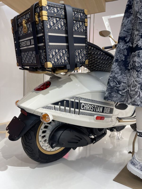 A white scooter with a large Christian Dior logo is parked indoors, featuring a patterned trunk on the back.