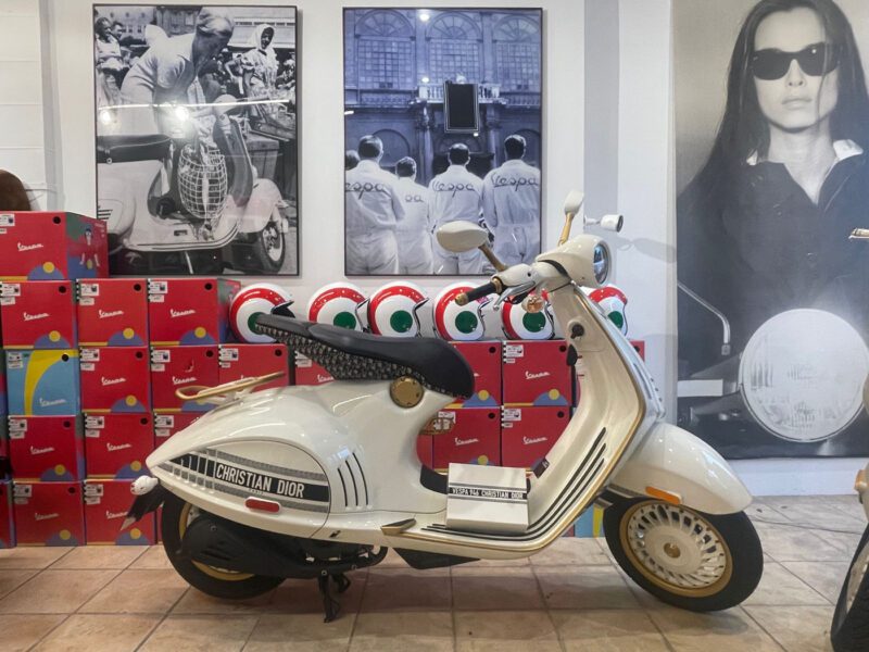 A white Christian Dior scooter is displayed indoors, surrounded by colorful helmets and graphic posters on the wall.