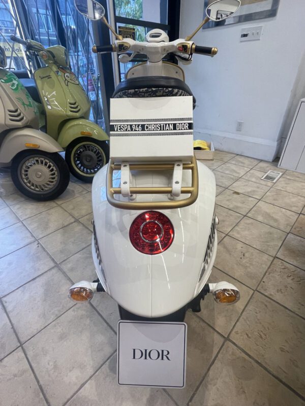 A white Vespa 946 Christian Dior scooter displayed indoors. The brand name "Dior" is prominently featured on a sign near the floor.