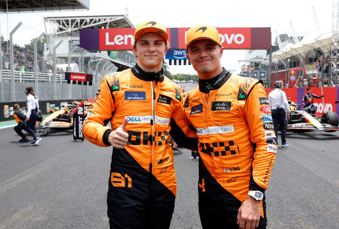 Two race car drivers in orange uniforms stand confidently on the starting grid, one giving a thumbs up as the F1 2025 season unfolds. Racing cars and a Lenovo-branded sign provide an exhilarating backdrop to this high-speed spectacle.