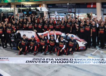 A racing team, comprised of accomplished works drivers, celebrates their FIA WEC 2024 World Endurance Championship win, posing triumphantly around a Porsche with arms raised in a cheering gesture.