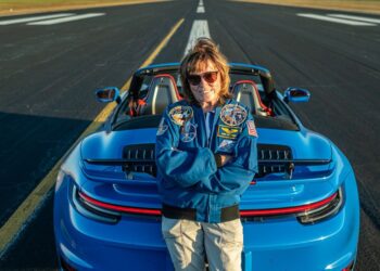 A person in a blue jacket and sunglasses stands with arms crossed, exuding confidence next to a blue convertible Porsche 911 Turbo parked on an airstrip, evoking the cool charisma of an astronaut prepping for launch.