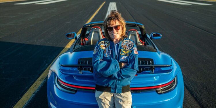 A person in a blue jacket and sunglasses stands with arms crossed, exuding confidence next to a blue convertible Porsche 911 Turbo parked on an airstrip, evoking the cool charisma of an astronaut prepping for launch.
