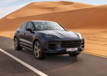 A dark blue Cayenne S Coupe cruises along a paved road, navigating the desert landscape with large sand dunes in the background, as part of an exhilarating Porsche Road Trip from Dubai to Abu Dhabi.