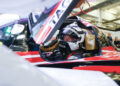 A race car driver, one of the esteemed Porsche Works Drivers, sits in a parked car, helmet and racing suit on. Engineers and team members buzz around in the background amidst the excitement of FIA WEC 2024 preparations.