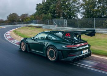 A black Porsche 911 GT3 RS, enhanced with a new Manthey Racing kit, zooms along a curved track surrounded by trees and guardrails, its large rear spoiler slicing effortlessly through the air.