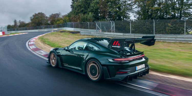 A black Porsche 911 GT3 RS, enhanced with a new Manthey Racing kit, zooms along a curved track surrounded by trees and guardrails, its large rear spoiler slicing effortlessly through the air.