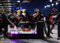 At the FIA WEC 2024, a Porsche racing car makes a pit stop at night. Crew members in red and black uniforms efficiently work on the vehicle, refueling and changing tires alongside their skilled Works Drivers.