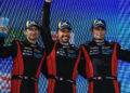 Three race car drivers in matching team uniforms celebrate on a podium, holding trophies. Wearing blue caps with yellow detailing, they stand against a backdrop featuring various logos from the 2024 Porsche FIA WEC season.