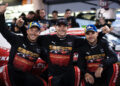 Three Porsche works drivers celebrate with arms around each other, wearing matching team uniforms and caps, standing proudly in front of their race car after a thrilling FIA WEC 2024 event.